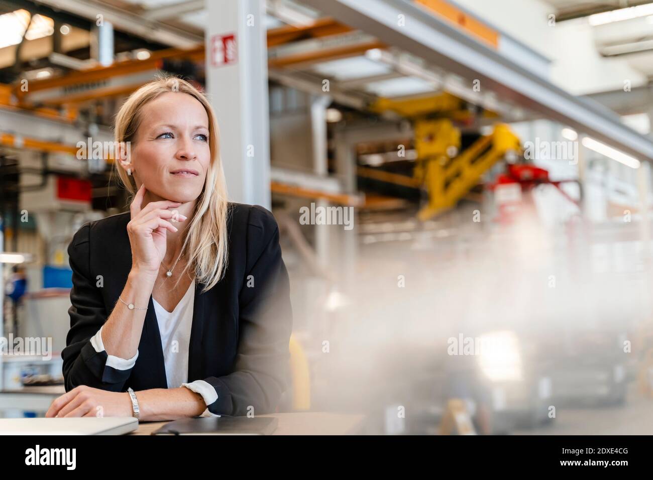 Femme d'affaires souriante et attentionnés, main sur le menton, assise en usine Banque D'Images