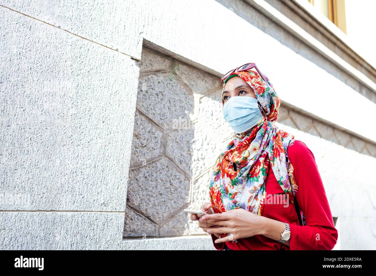 Femme en hijab portant un masque de protection tenant un smartphone Tout en marchant par le mur pendant COVID-19 Banque D'Images