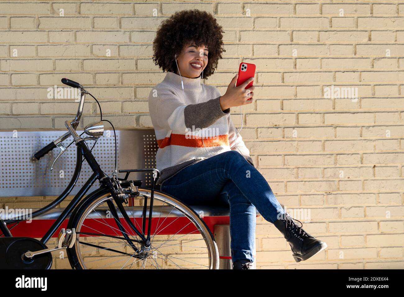 Une femme heureuse prenant le selfie tout en étant assise sur le banc à vélo contre le mur de briques Banque D'Images