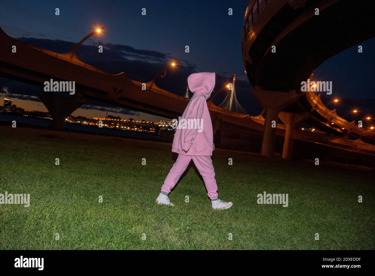 Femme portant un costume de course rose et une capuche marchant sur la pelouse la nuit, sous les autoroutes surélevées Banque D'Images