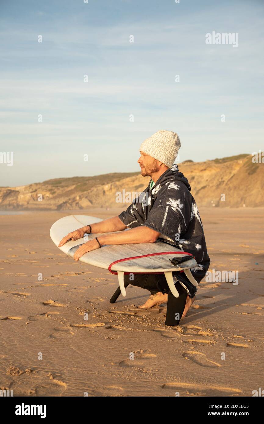 Un surfeur mâle qui regarde loin tout en s'accroupiant avec planche de surf à la plage Banque D'Images