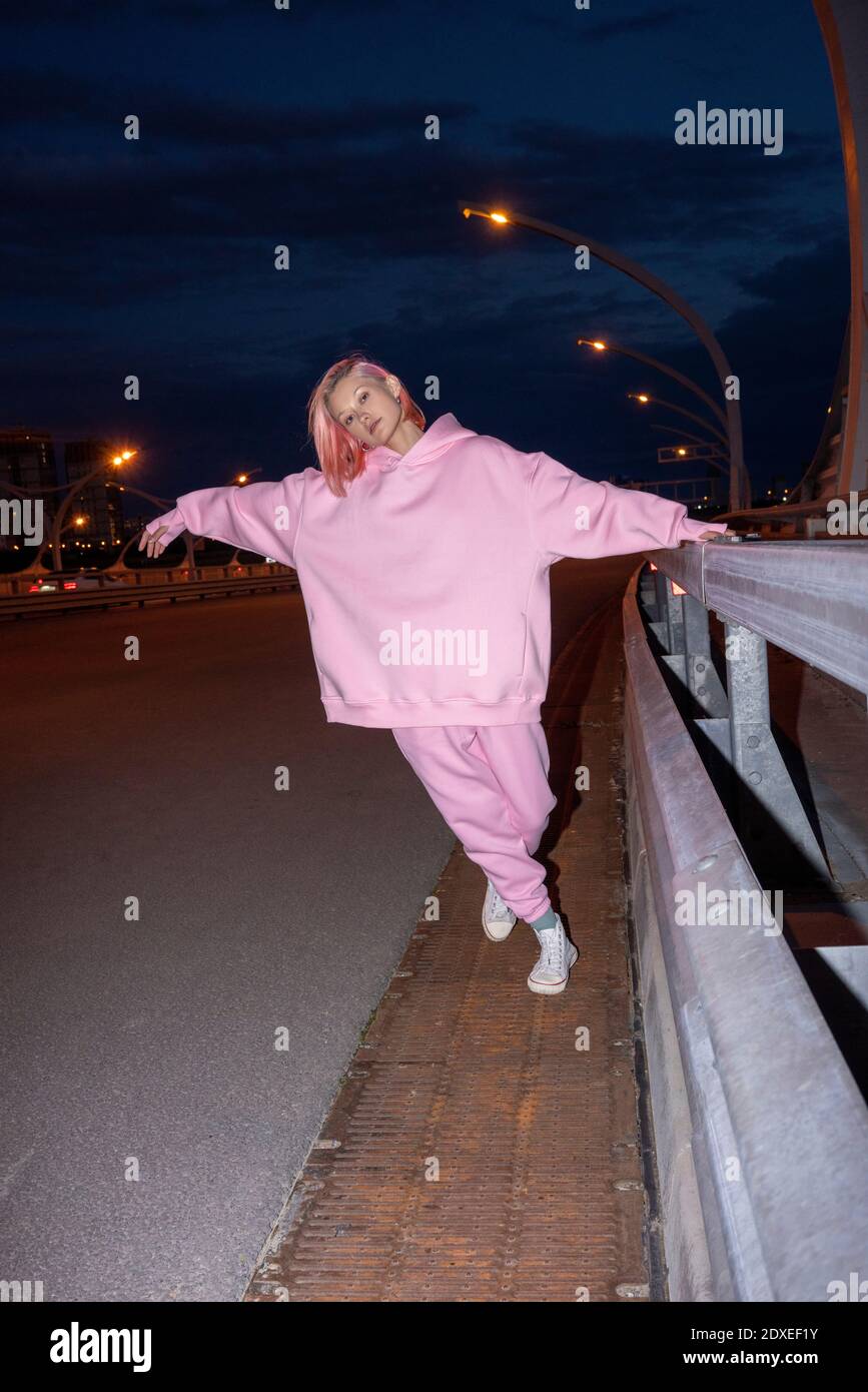 Jeune femme aux cheveux roses portant un costume d'athlétisme rose debout à la barrière routière la nuit Banque D'Images