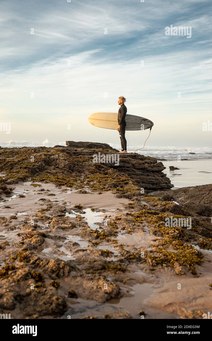 Surfeur mâle debout sur la formation rocheuse avec planche de surf à la plage Banque D'Images