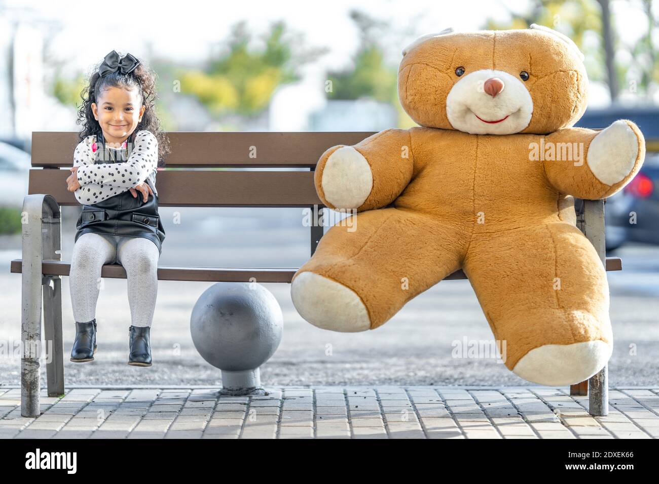 Fille souriante assise avec les bras croisés par l'ours en peluche à distance sociale sur banc Banque D'Images