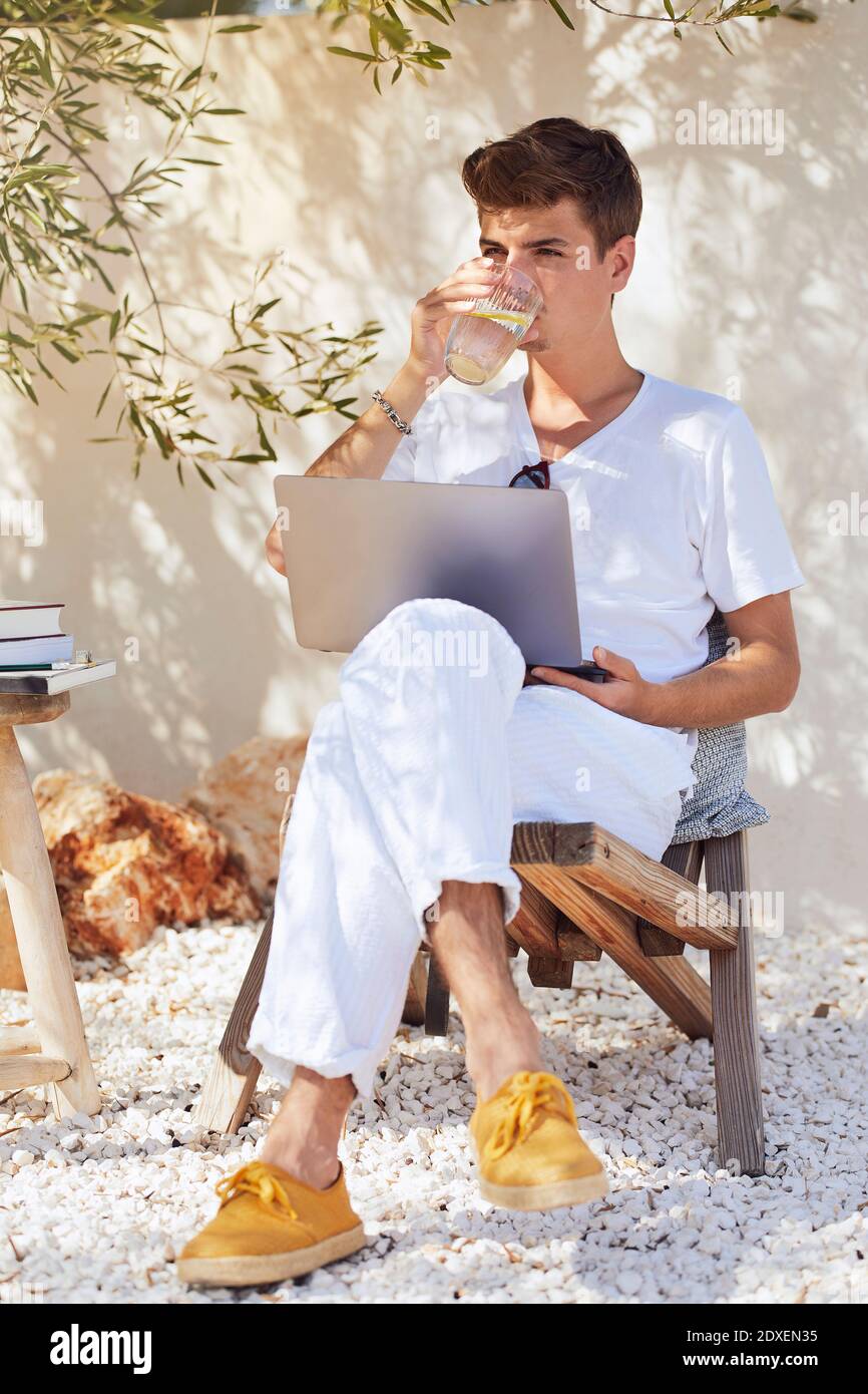 Jeune homme avec de l'eau potable pour ordinateur portable tout en étant assis sur une chaise contre le mur Banque D'Images
