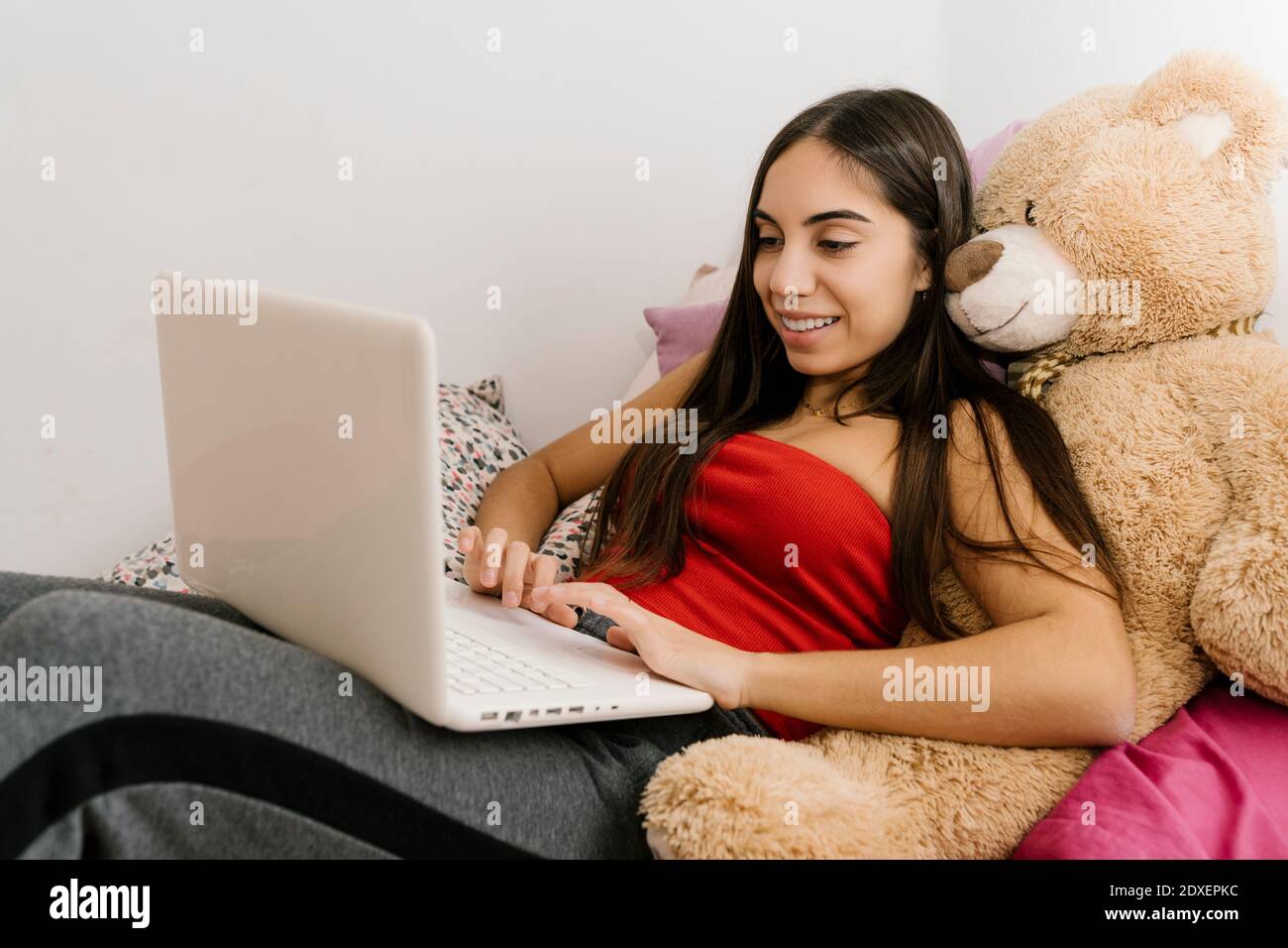Adolescente souriante utilisant un ordinateur portable tout en se penchant sur l'ours en peluche jouet à la maison Banque D'Images