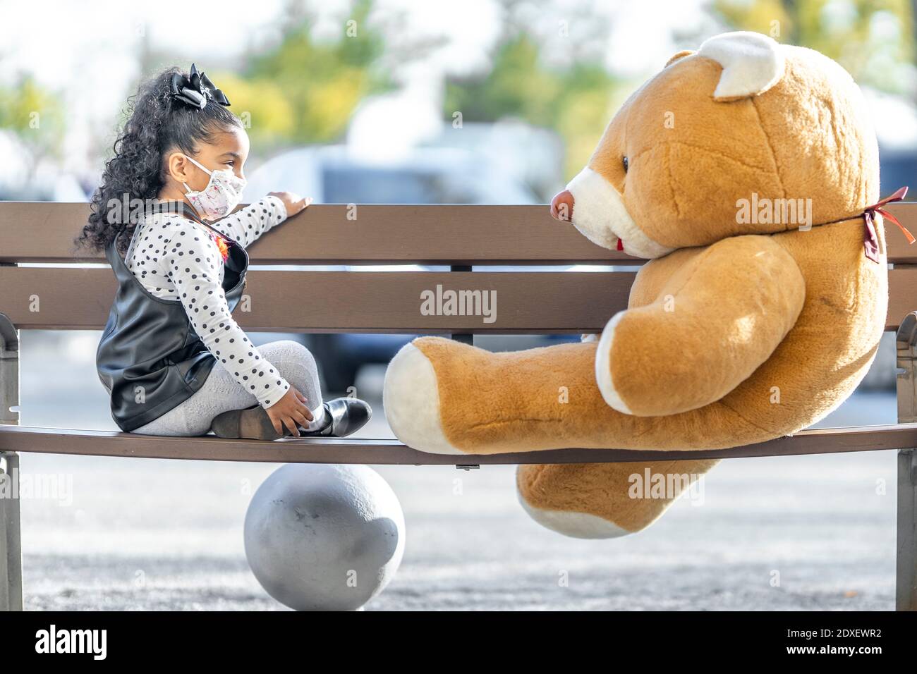 Fille portant un masque facial jouant avec l'ours en peluche pendant qu'elle est assise sur banc Banque D'Images