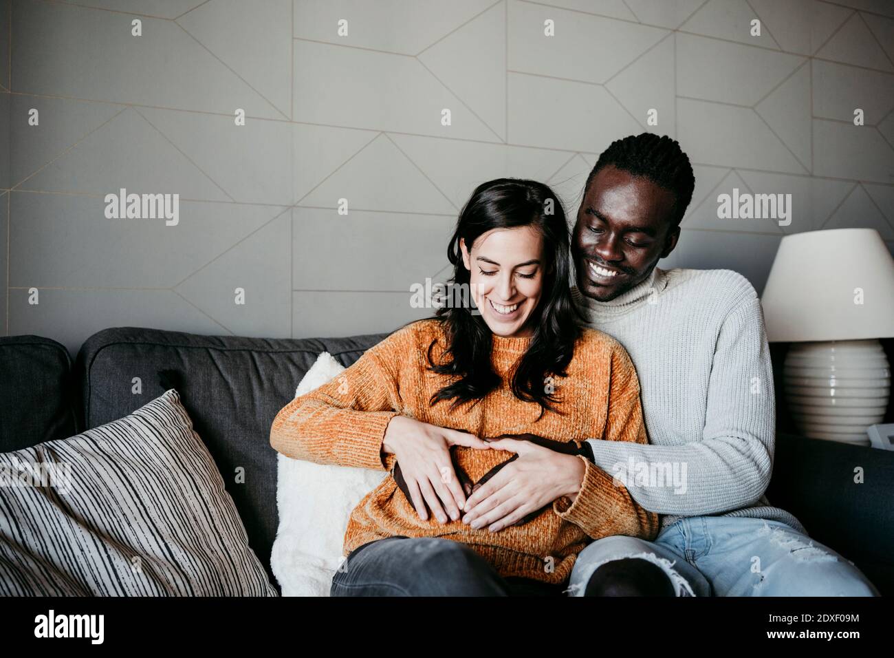 Homme heureux et femme enceinte formant le coeur avec les mains sur le ventre tout en étant assis sur un canapé à la maison Banque D'Images