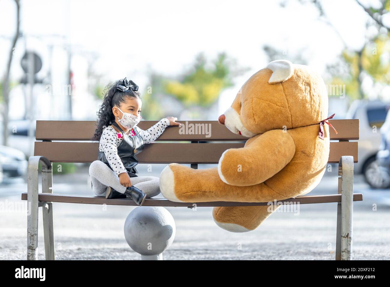Fille portant un masque facial jouant avec l'ours en peluche pendant qu'elle est assise à distance sociale sur banc Banque D'Images