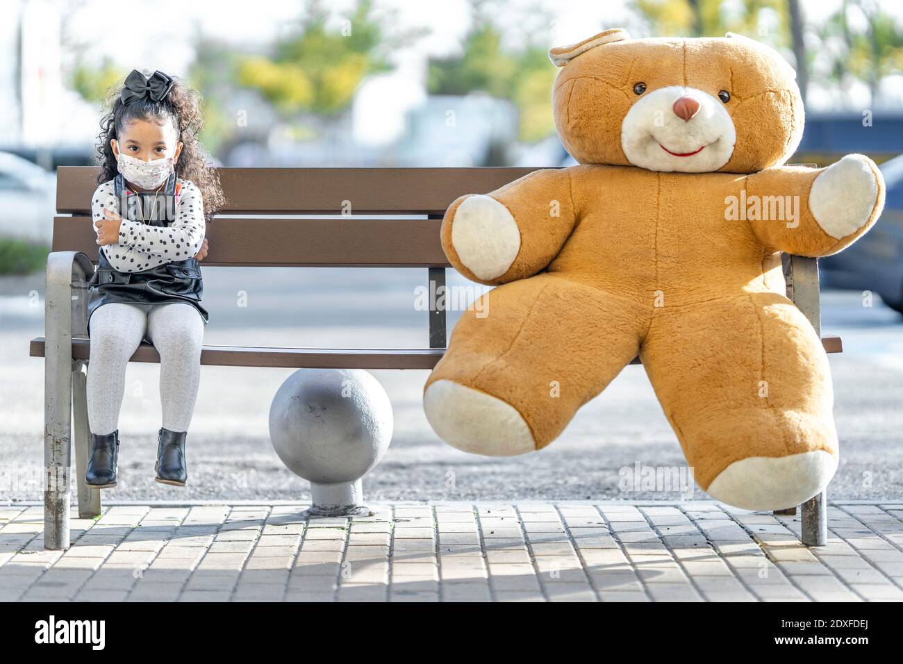 Fille portant un masque de visage assis avec un ours en peluche à la vie sociale distance sur banc Banque D'Images