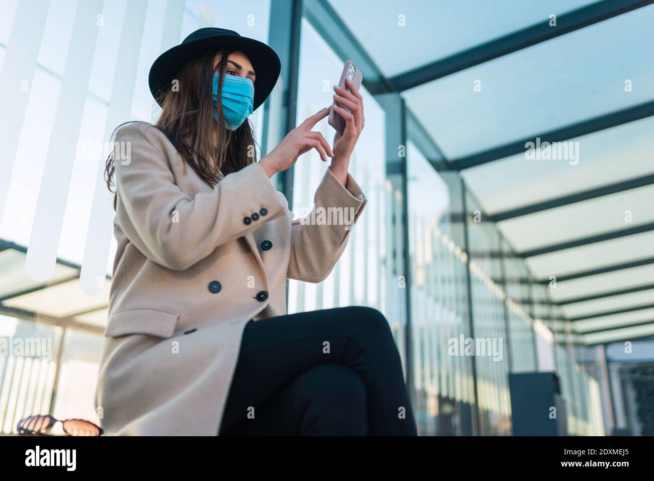 Fille dans le masque de protection est assis à l'arrêt de bus avec le téléphone dans les mains. La femme est assise à l'arrêt d'autobus pendant une pandémie et attend le transport Banque D'Images