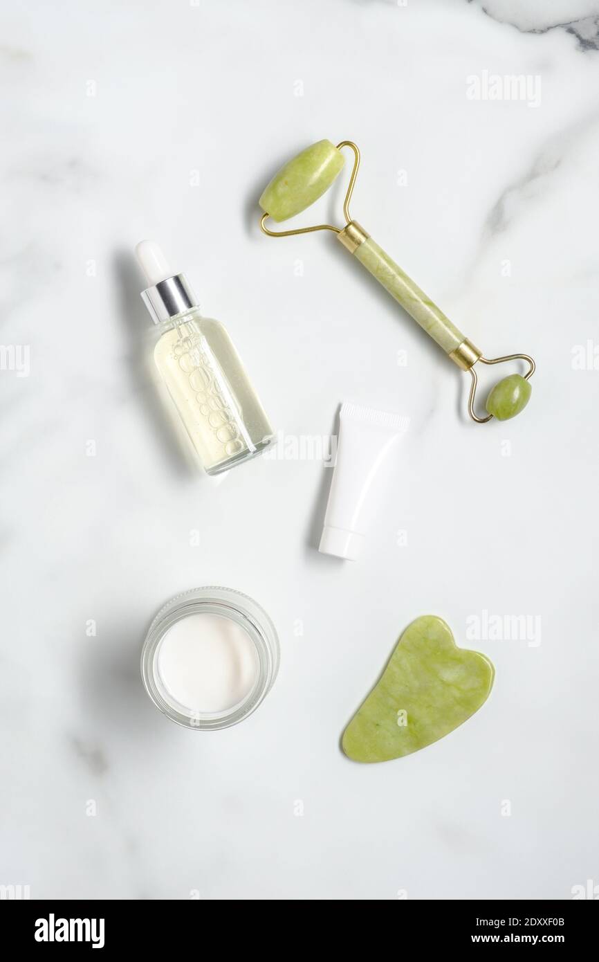 Rouleau facial en pierre de Jade, SHA gua, lotion de sérum, hydratant, tube avec crème sur le bureau en marbre. Flat lay, vue de dessus. Ensemble de cosmétiques anti-âge. Banque D'Images