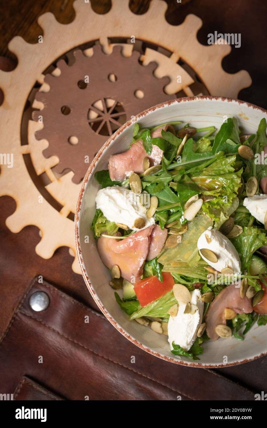 Bol de salade de légumes au saumon, fromage mozzarella sur une table recouverte d'un chiffon en cuir et un détail décoratif d'un mécanisme simple Banque D'Images