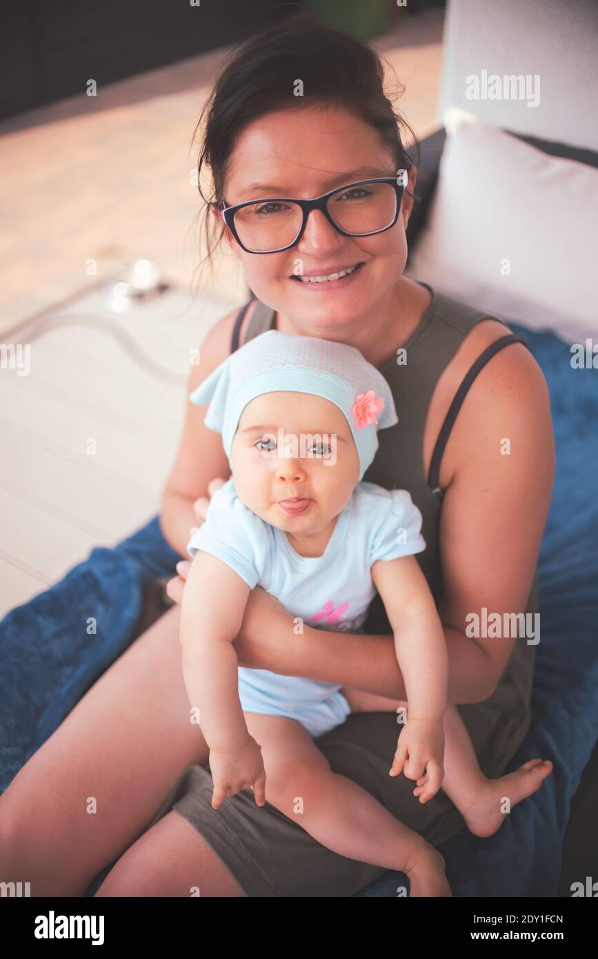 Bébé avec l'expression drôle et la langue dehors est assis sur les genoux de mère. Banque D'Images