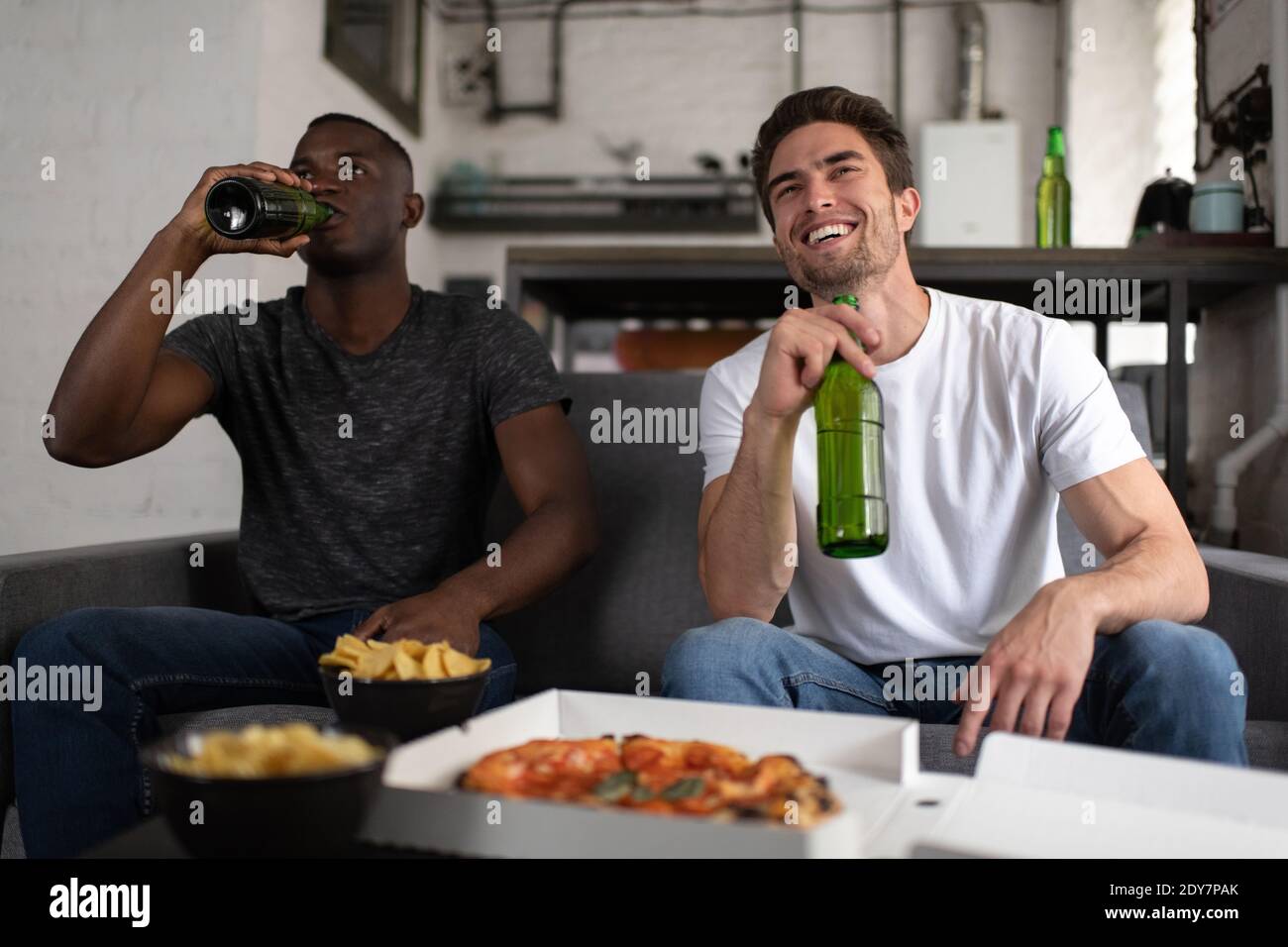 Heureux hommes multiethniques dans des tenues décontractées boire de la bière froide et souriant assis sur un canapé près de la table avec des en-cas et regarder le match de football toget Banque D'Images