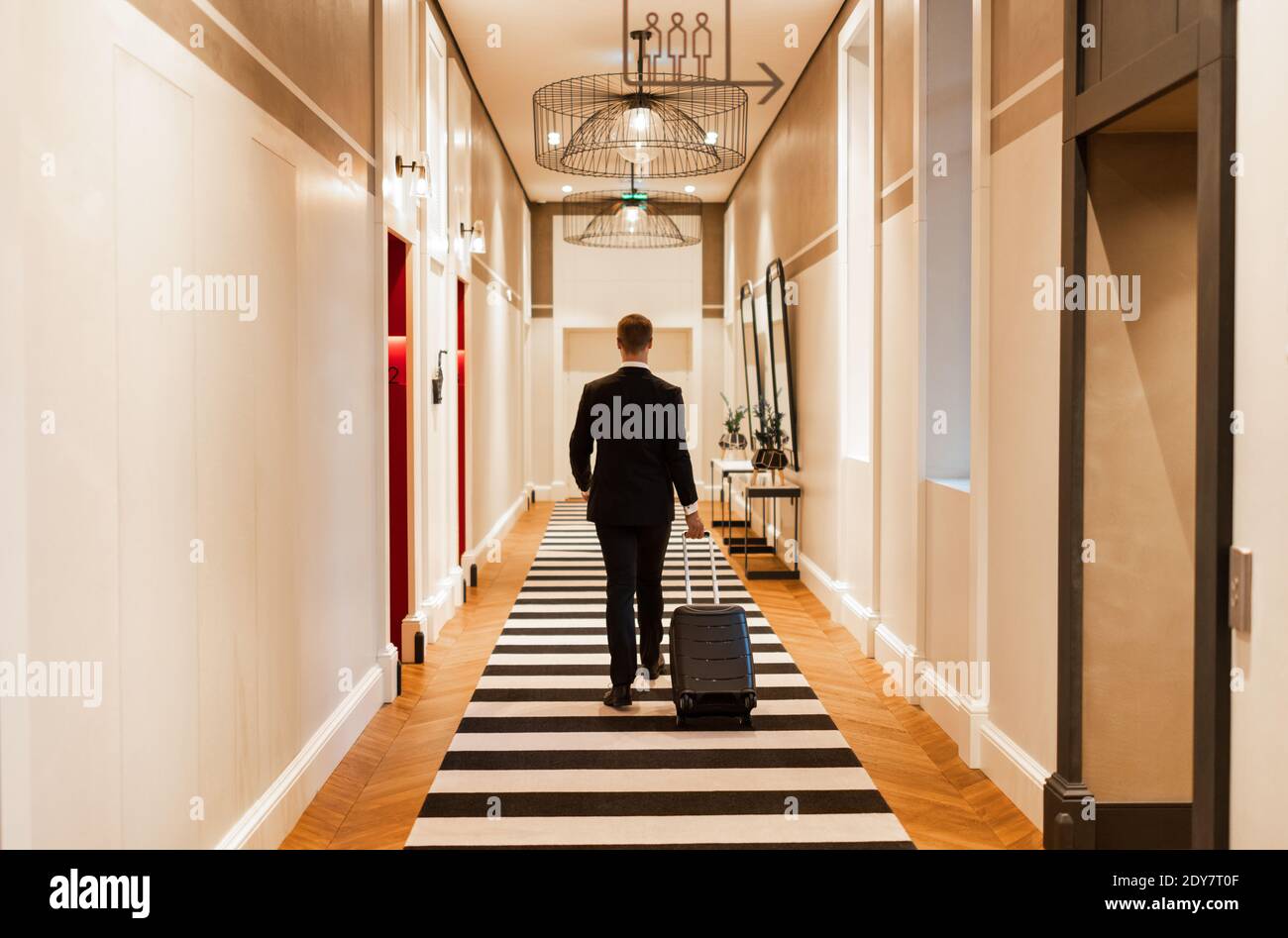 Vue arrière de l'homme adulte en costume noir tirant la valise tout en marchant dans le couloir lumineux de l'hôtel de luxe pendant voyage d'affaires Banque D'Images