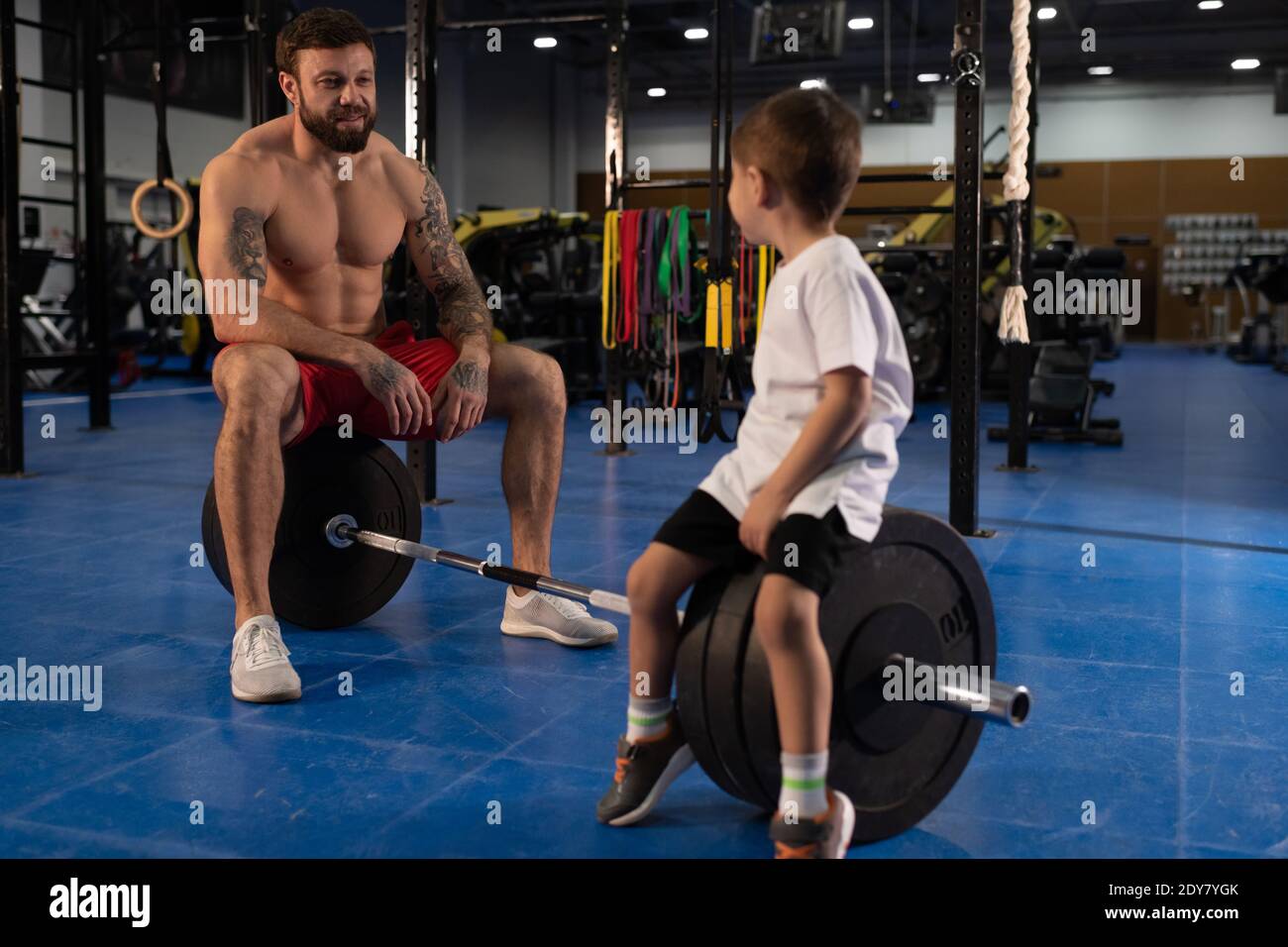Athlète masculin fort et petit garçon assis sur barbell et se regardant les uns les autres tout en prenant le repos pendant l'entraînement d'haltérophilie dans la salle de sport Banque D'Images