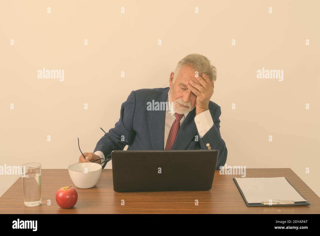 Photo de studio d'un homme d'affaires barbu de haut niveau tenant des lunettes de vue pendant avoir des maux de tête avec un ordinateur portable et des choses de base pour le travail table en bois Banque D'Images