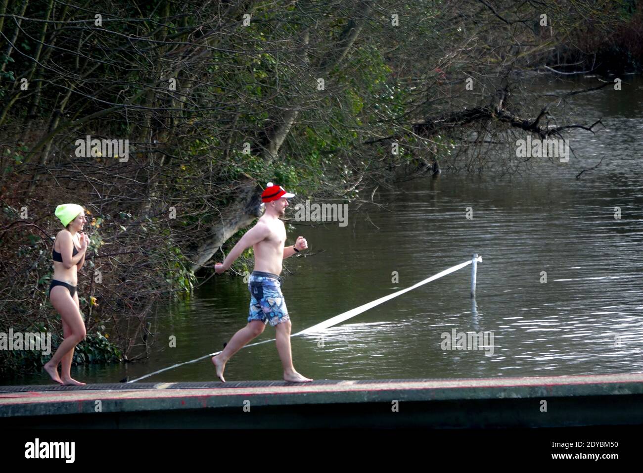 Deux nageurs descendent la jetée Hampstead Men's Pond pour un événement annuel de natation de Noël le jour de Noël 2020. Un homme portant un chapeau de père noël sautant la jetée, suivi d'une jeune femme qui frite le maillot. Banque D'Images