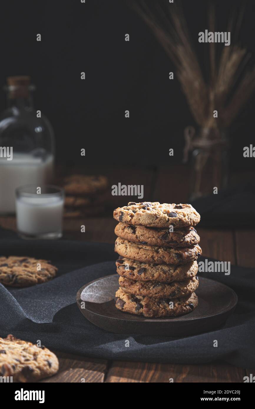 Biscuits au chocolat empilés, avec un verre et une bouteille de lait sur une base en bois, fond sombre. Concept de nourriture douce. Banque D'Images