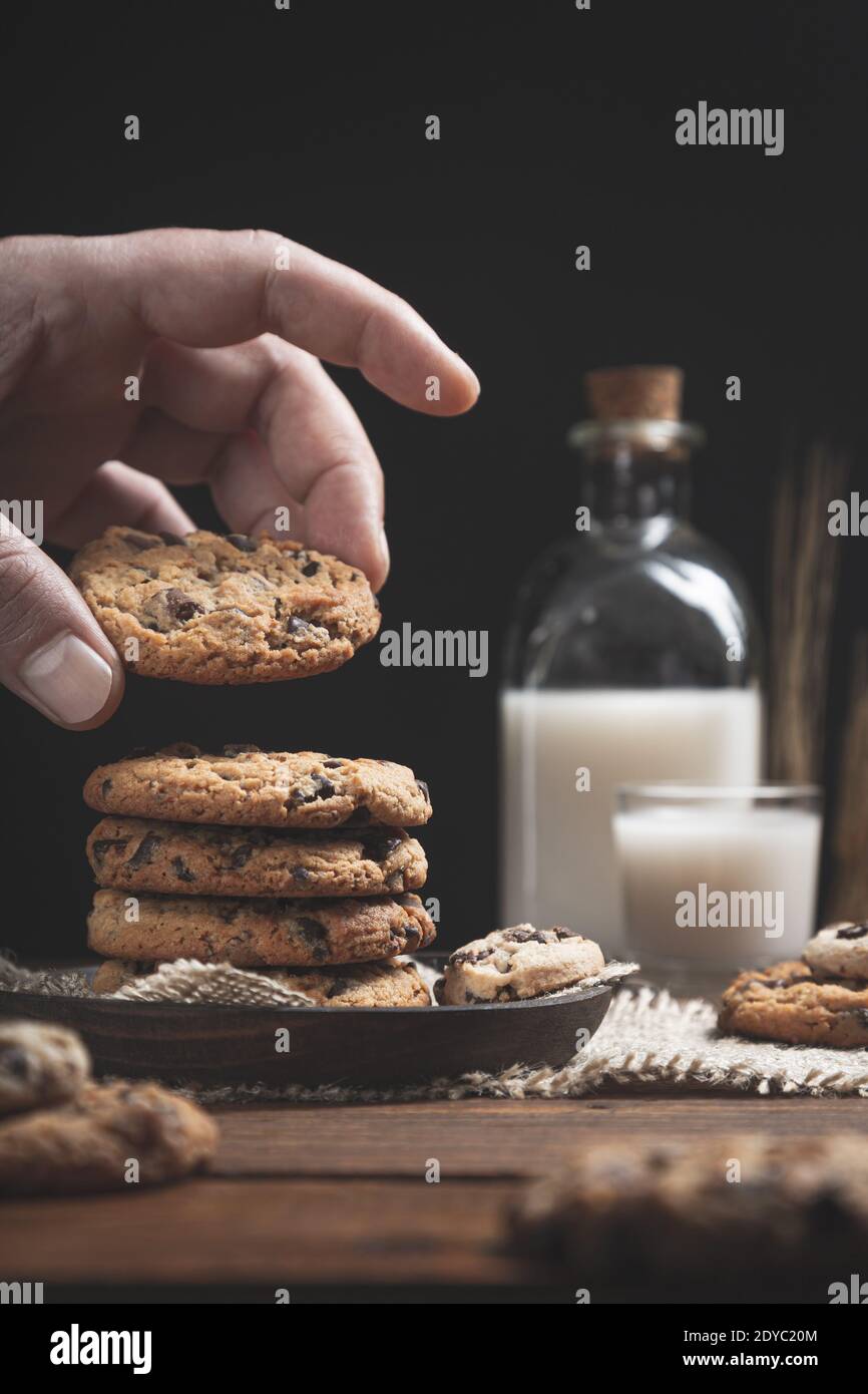 Main tenant un biscuit de pépites de chocolat avec un verre et une bouteille de lait sur une base en bois, fond sombre. Concept de nourriture douce. Banque D'Images