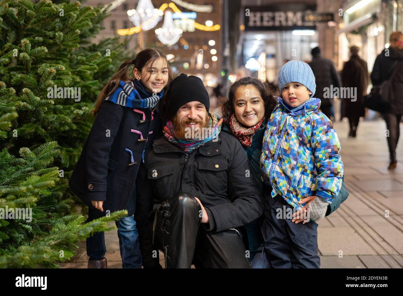 Bonne famille multinationale avec maman, papa et enfants à l'extérieur dans un centre-ville à l'heure de Noël. En train de rouler dans l'appareil photo Banque D'Images