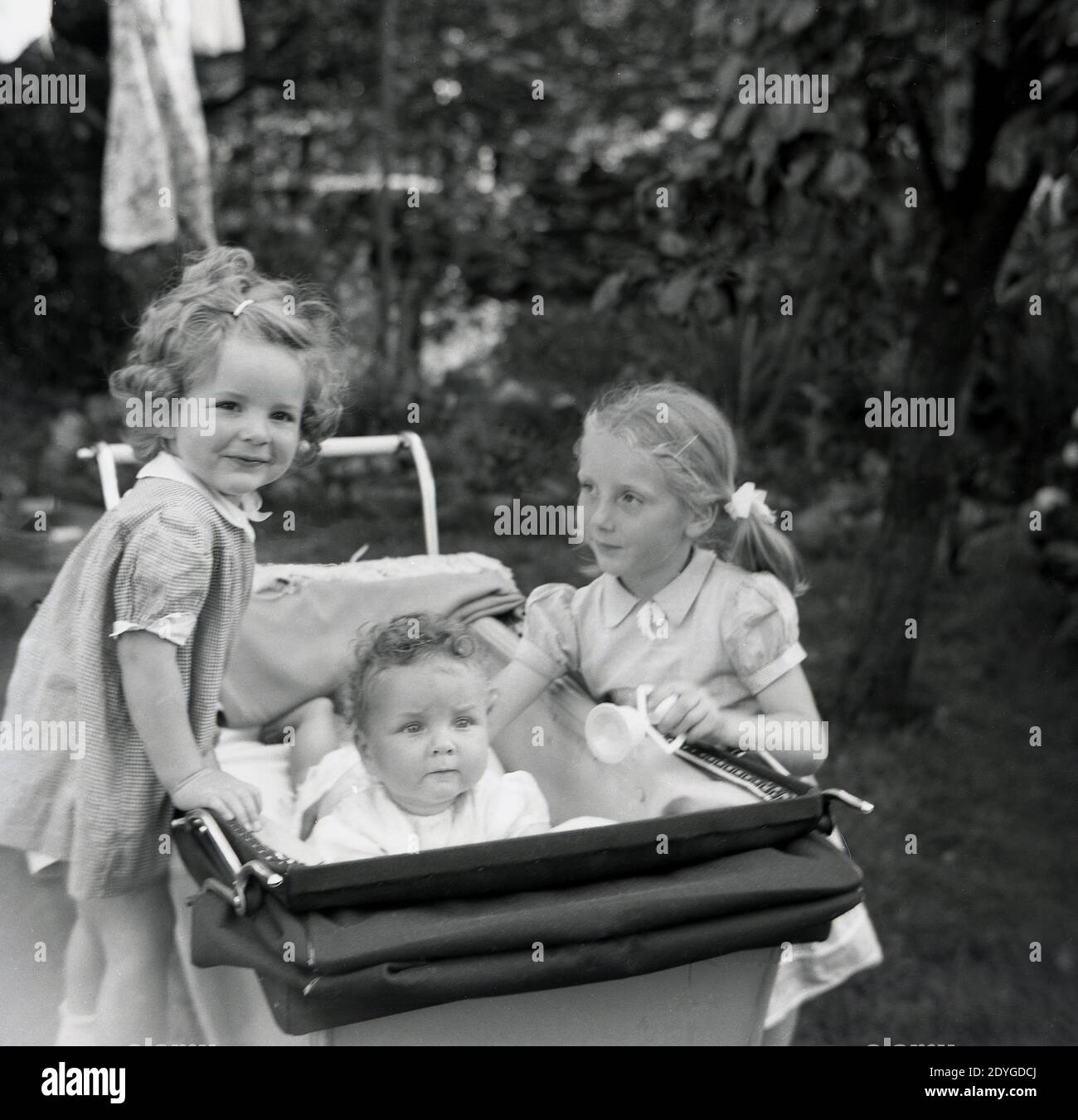 Années 1950, historique, l'été et à l'extérieur dans un jardin, deux petites filles douces, sœurs, debout à côté d'un entraîneur-construit pram ou de la voiture de bébé de l'époque, avec l'une de la fille debout sur la roue et l'autre avec sa main sur leur bébé frère couché dans le pram, Angleterre, Royaume-Uni. Banque D'Images