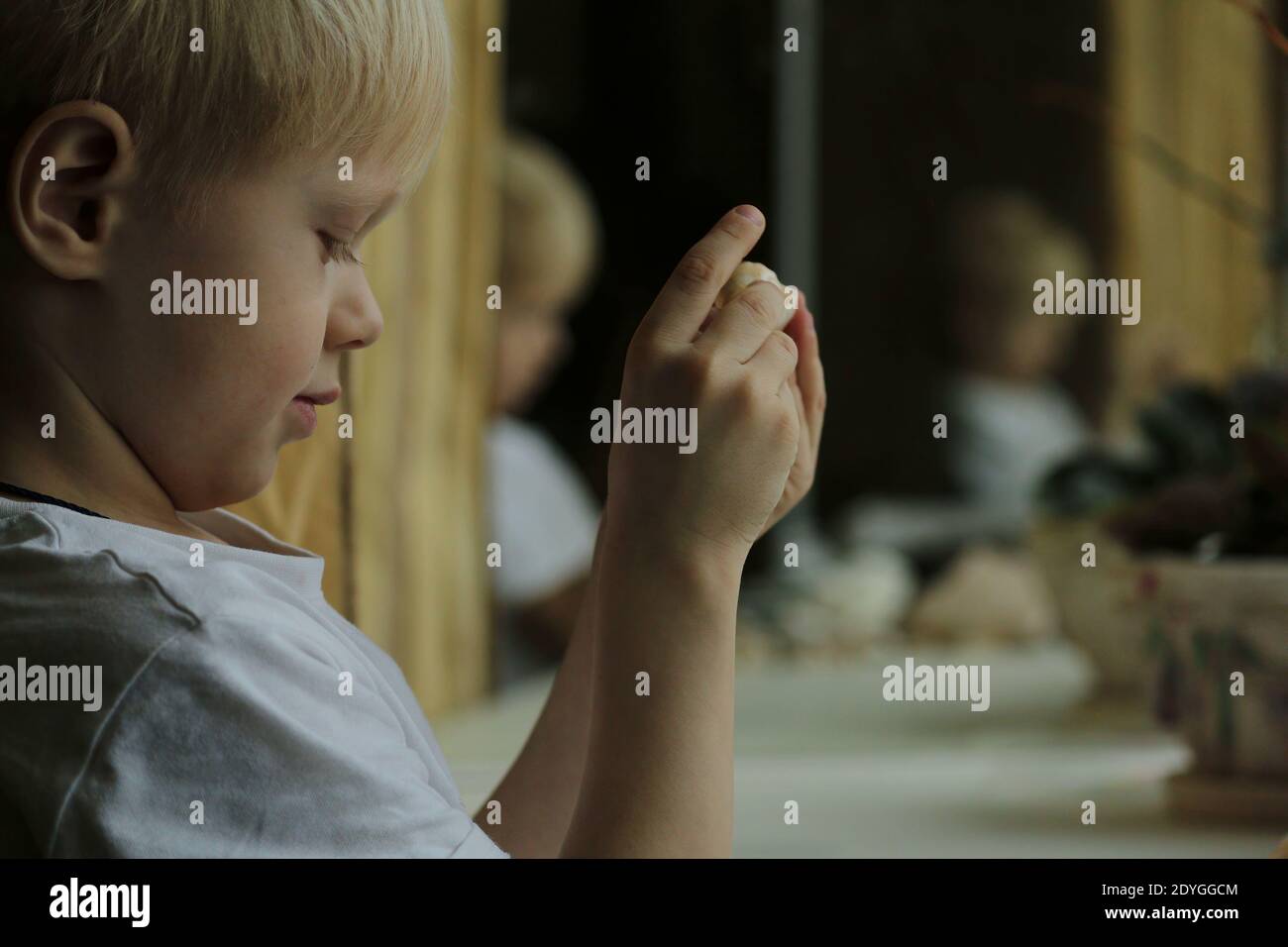 Un petit garçon aux cheveux blonds dans un T-shirt blanc se tient en profil par une fenêtre intérieure, reflétée dans les miroirs, et tient des coquillages dans ses mains Banque D'Images