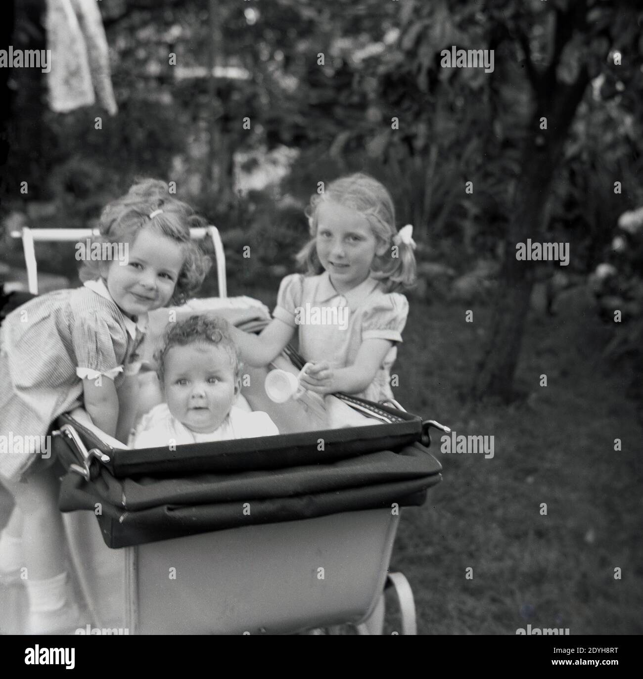 Années 1950, historique, à l'extérieur dans un jardin, deux jeunes filles debout à côté d'un landau construit par autocar ou d'une voiture d'enfant de l'époque avec leur petite sœur à l'intérieur, avec la petite fille debout sur le volant, Angleterre, Royaume-Uni. Banque D'Images