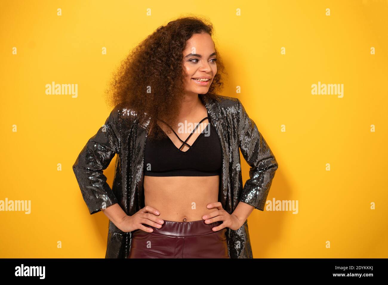 Bonne fille afro-américaine avec belle coiffure posant le sourire regardant côté loin avec la main sur les hanches portant blouson noir brillant et haut noir. Heureux Banque D'Images