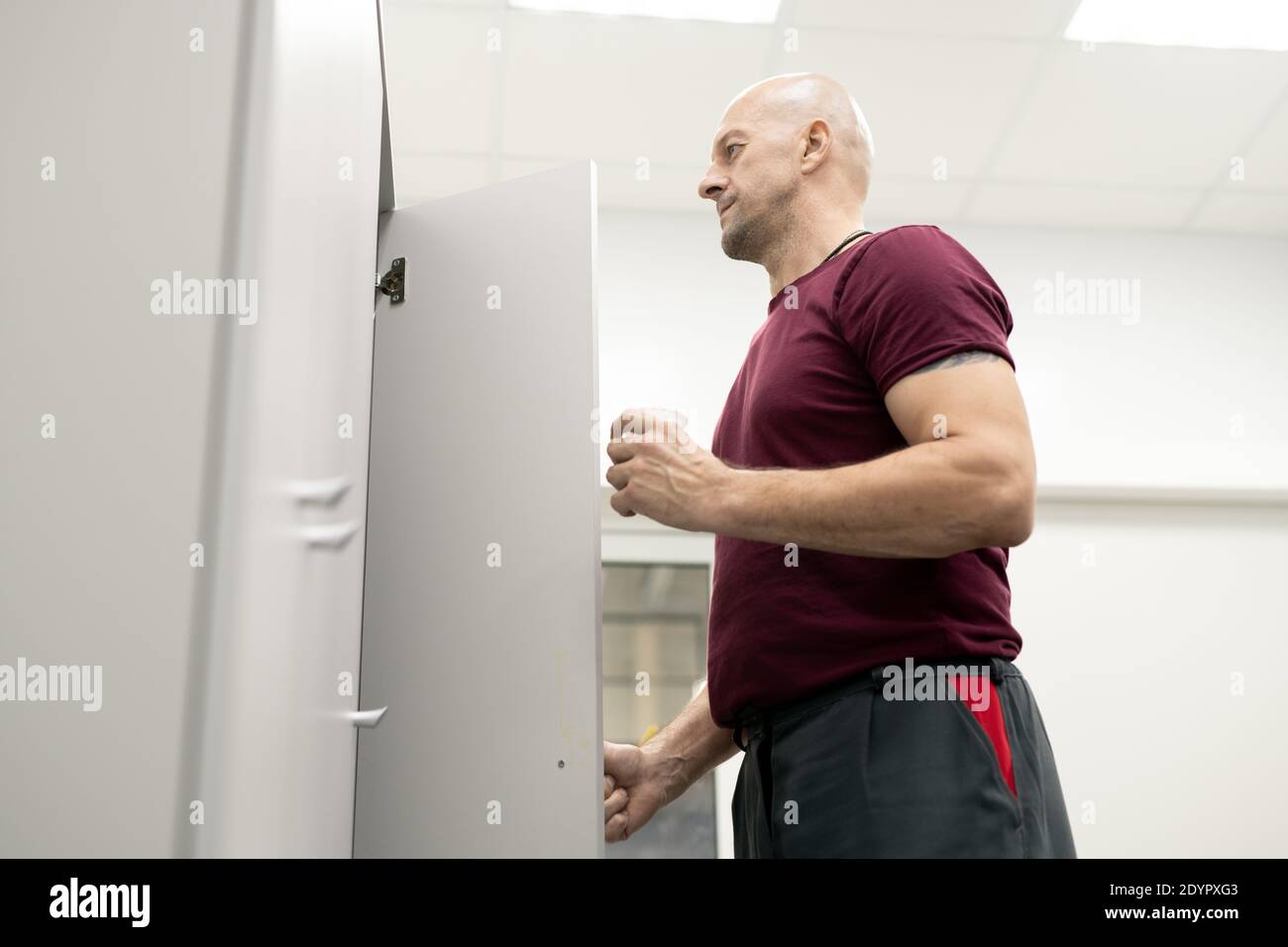Vue latérale d'un ouvrier sérieux et sérieux de l'usine moderne boire tout en ouvrant la porte du casier avec ses vêtements de travail et d'autres choses Banque D'Images
