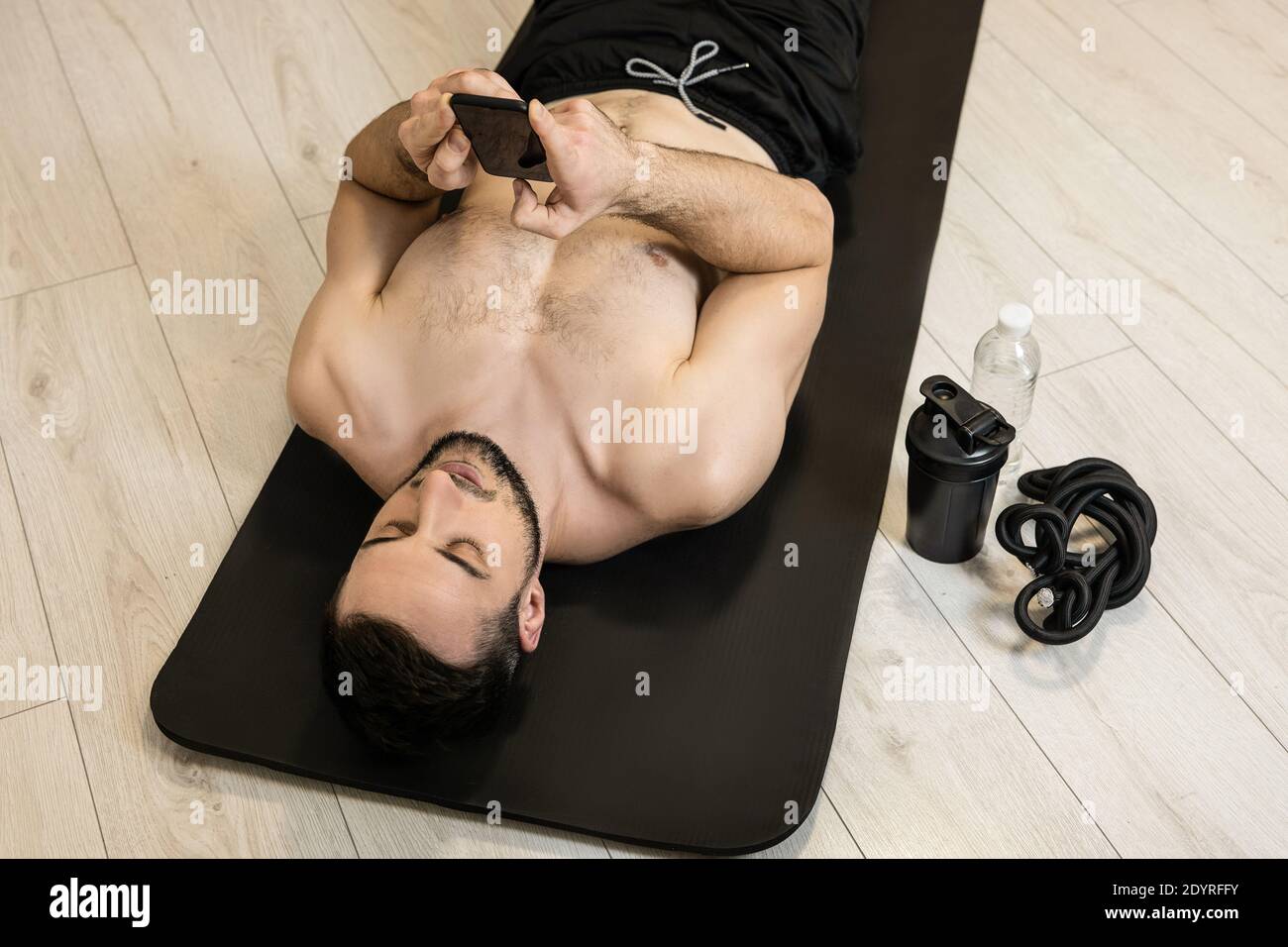 FIT MAN se repose après une forme physique dure et envoie des SMS avec des amis au téléphone. Équipements sportifs. Temps de repos. Paresseux allongé sur un tapis de yoga noir et Banque D'Images