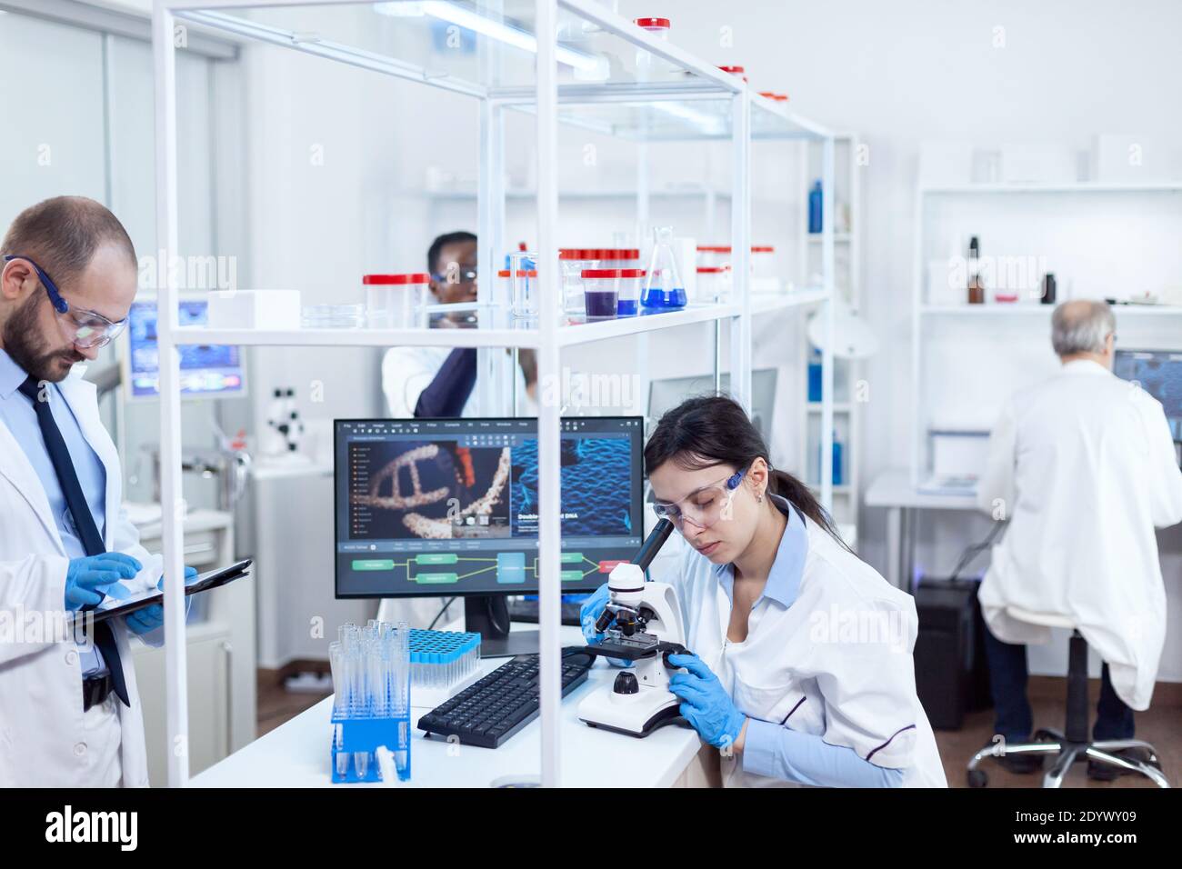 Scientifique utilisant le microscope pour l'étude de la médecine. Équipe de chercheurs en génie de la pharmacologie dans un laboratoire stérile pour l'industrie de la santé avec un assistant africain en arrière-plan. Banque D'Images