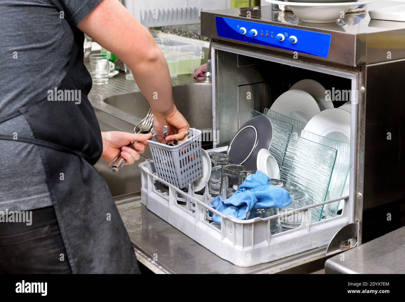 Ouvrier de cuisine de restaurant vider un lave-vaisselle enlever les couverts propres de le panier en gros plan sur ses mains et la porte ouverte de la machine Banque D'Images