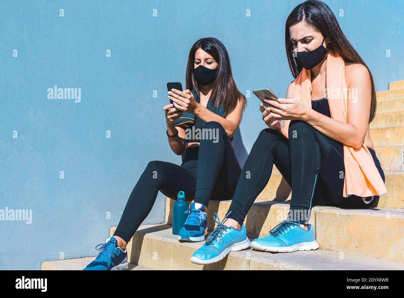 deux jeunes femmes portant des vêtements de sport et des masques noirs, assis sur des marches, faisant une pause et vérifiant leur smartphone. Concept de sport Banque D'Images