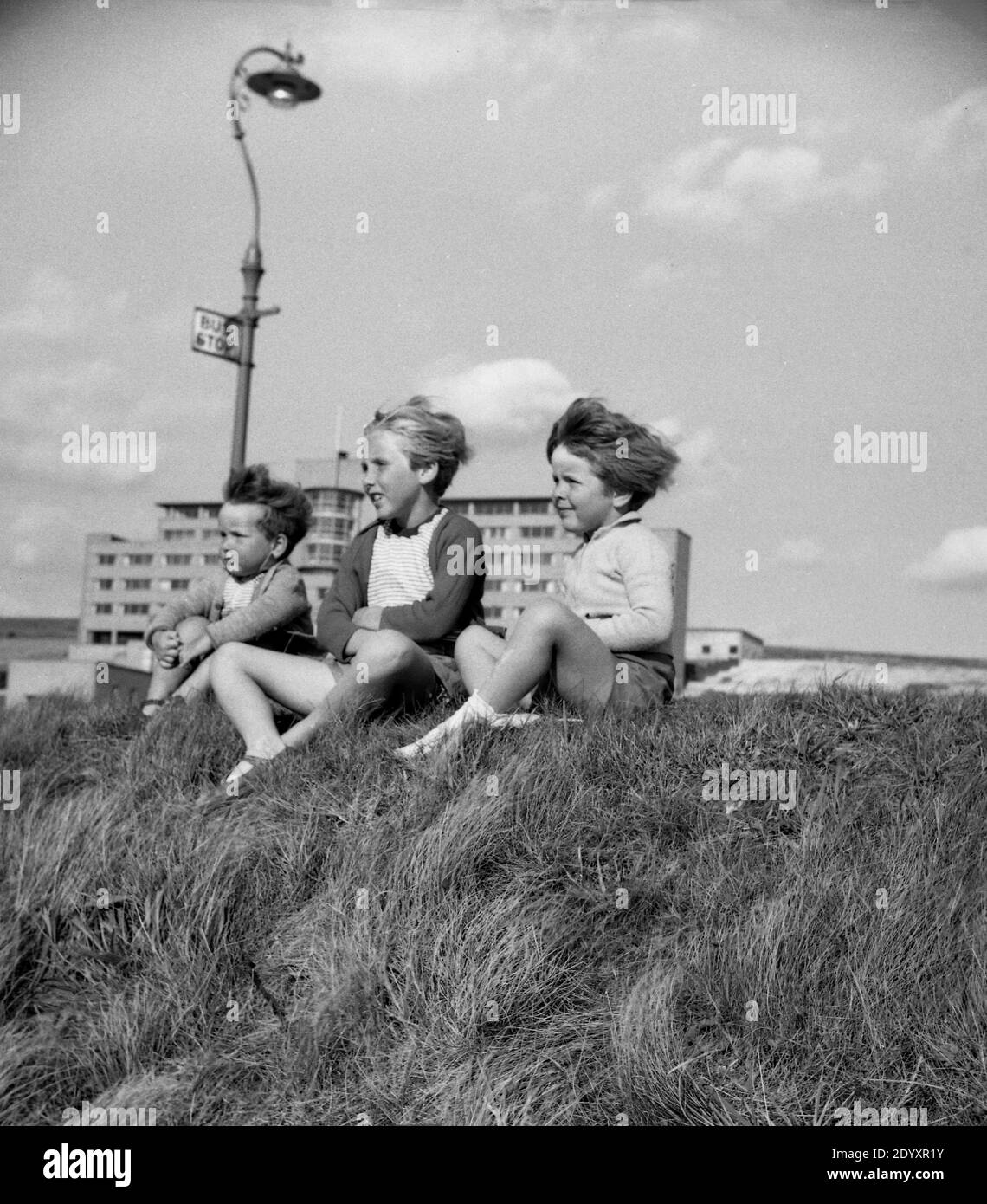 années 1950, historique, trois jeunes enfants assis ensemble sur une rive herbeuse à la côte regardant vers la mer. Banque D'Images