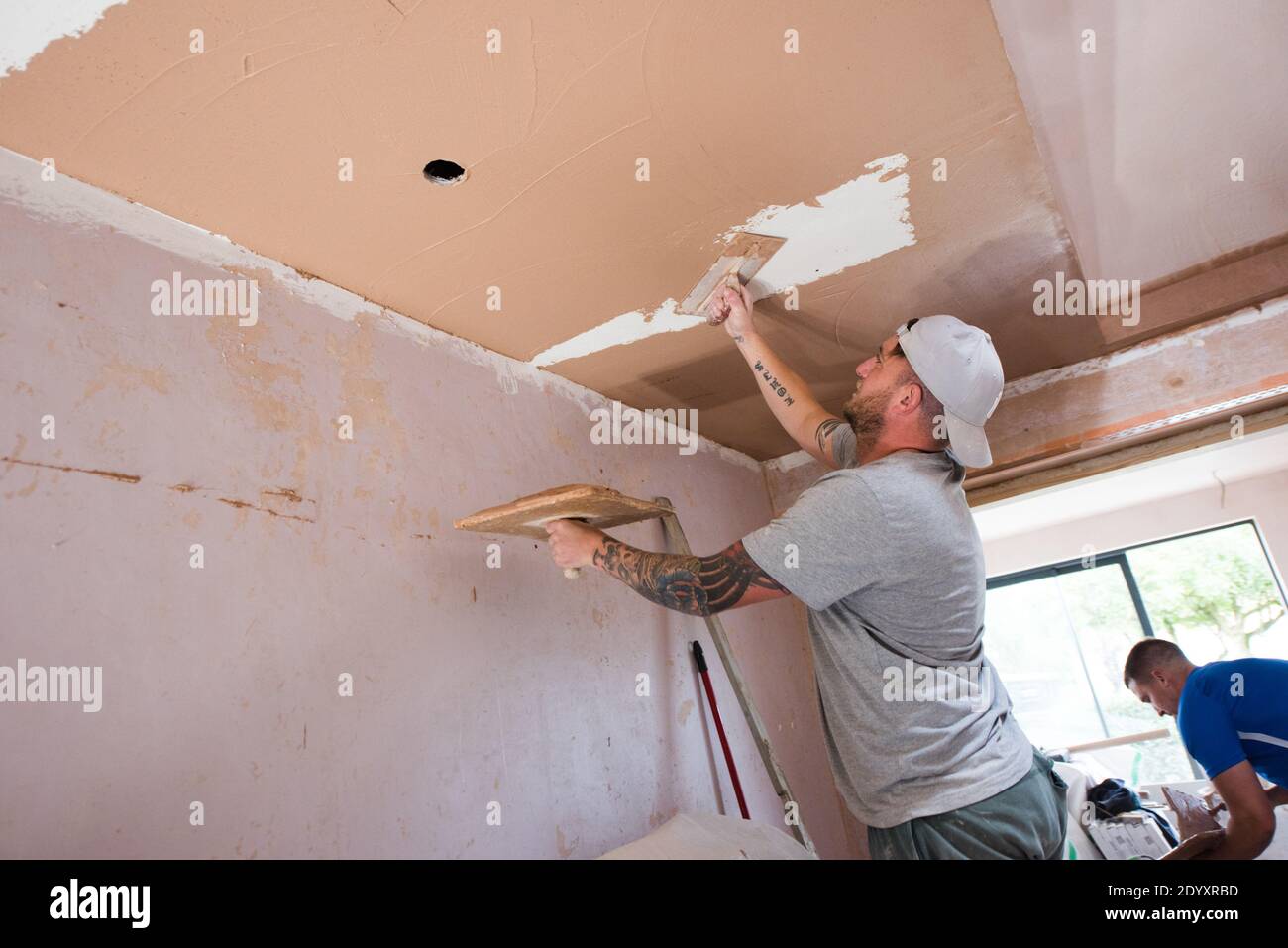 Homme blanc tatoué portant une casquette de baseball plâtrant les murs et plafond sur un projet de rénovation de maison Banque D'Images