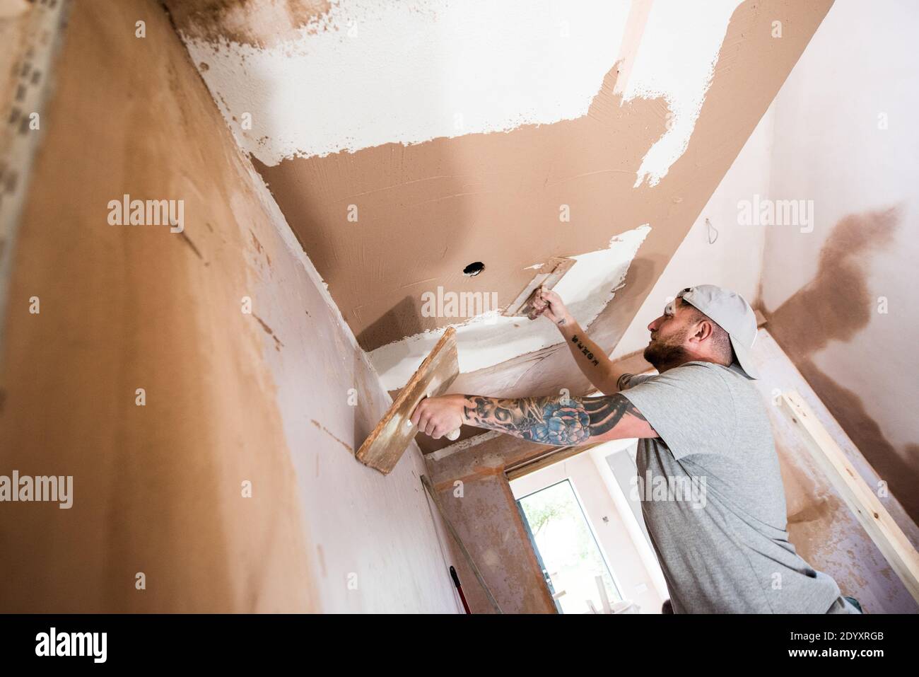 Homme blanc tatoué portant une casquette de baseball plâtrant les murs et plafond sur un projet de rénovation de maison Banque D'Images
