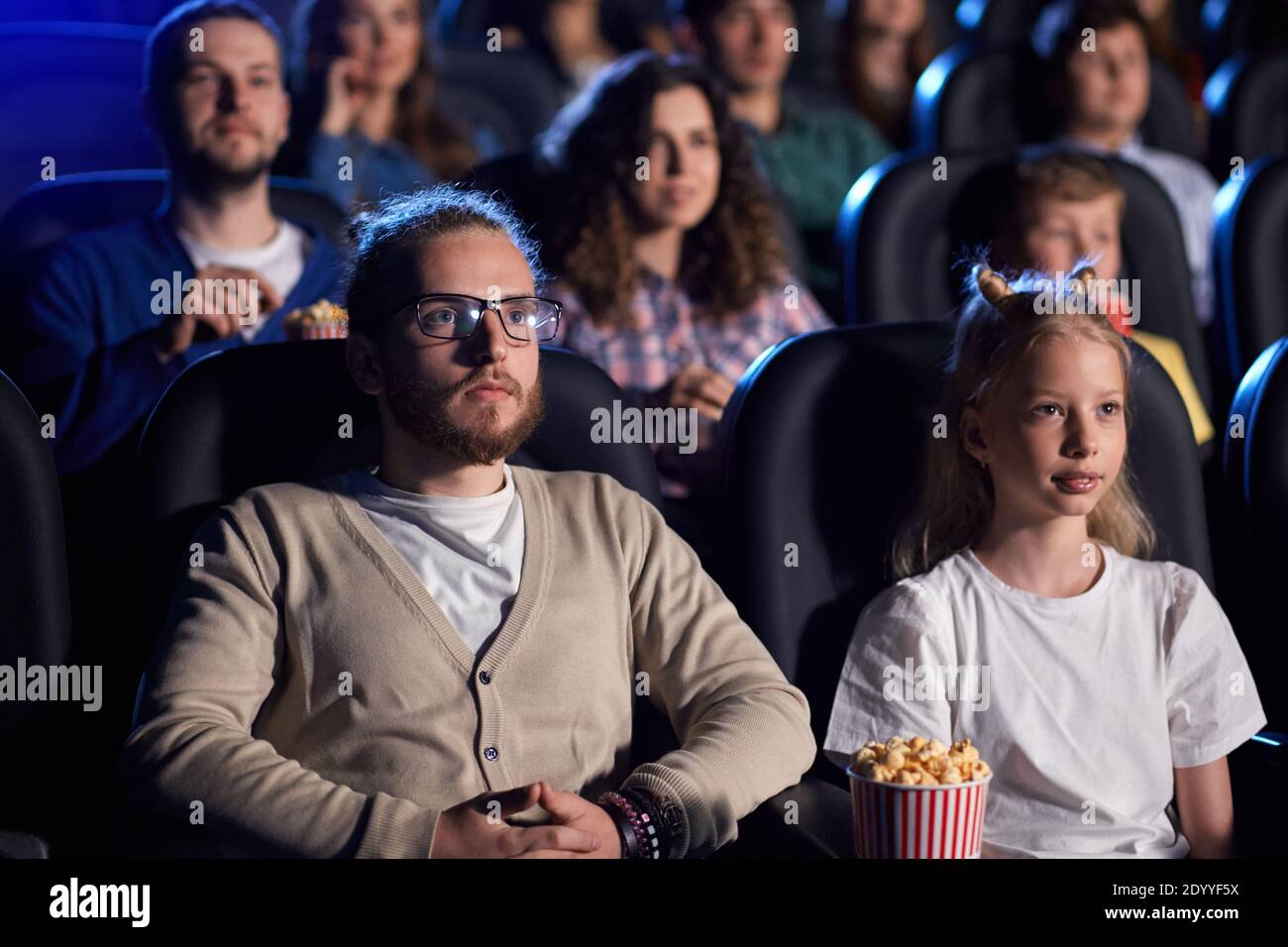 Vue latérale d'un beau barbu en lunettes regardant un film sérieux avec une jeune sœur blonde au cinéma, des visages forts. Adolescente auscasienne concentrée, en train de regarder un film d'horreur avec sa famille. Banque D'Images