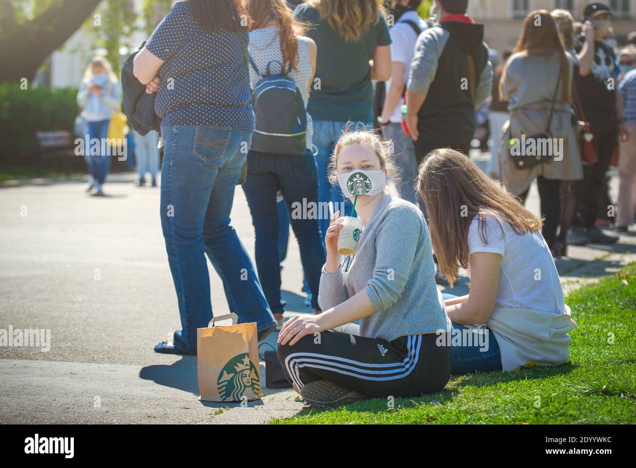 Une fille qui boit du café Starbucks sous un masque facial en tissu avec le logo Starbucks, est assise sur le sol à côté d'une file d'attente pour un test de virus corona. Banque D'Images