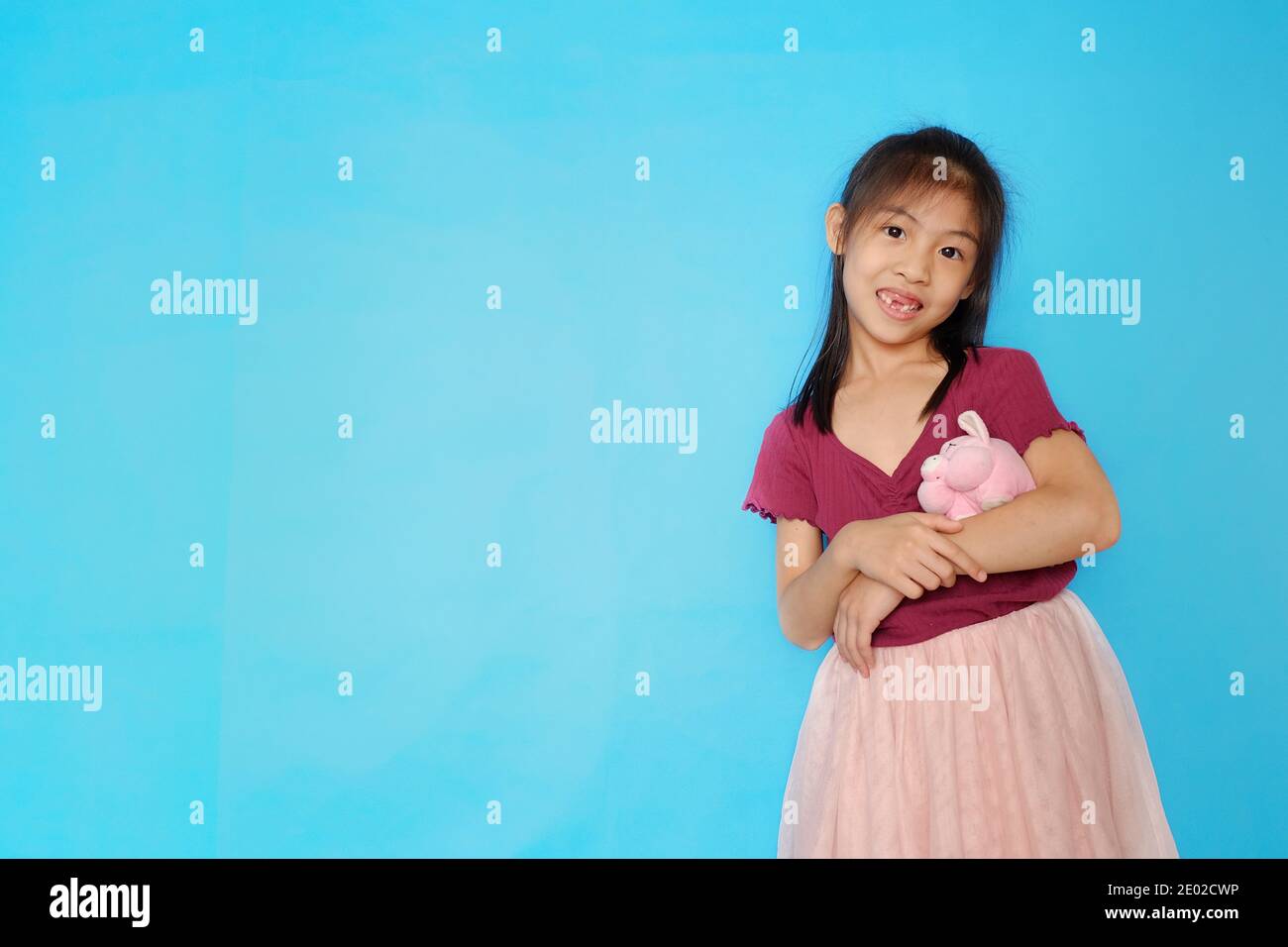Une photo de gros plan d'une jolie fille asiatique, regardant directement l'appareil photo, souriant, bertant sa poupée de porc rose avec fond bleu clair Uni. Banque D'Images
