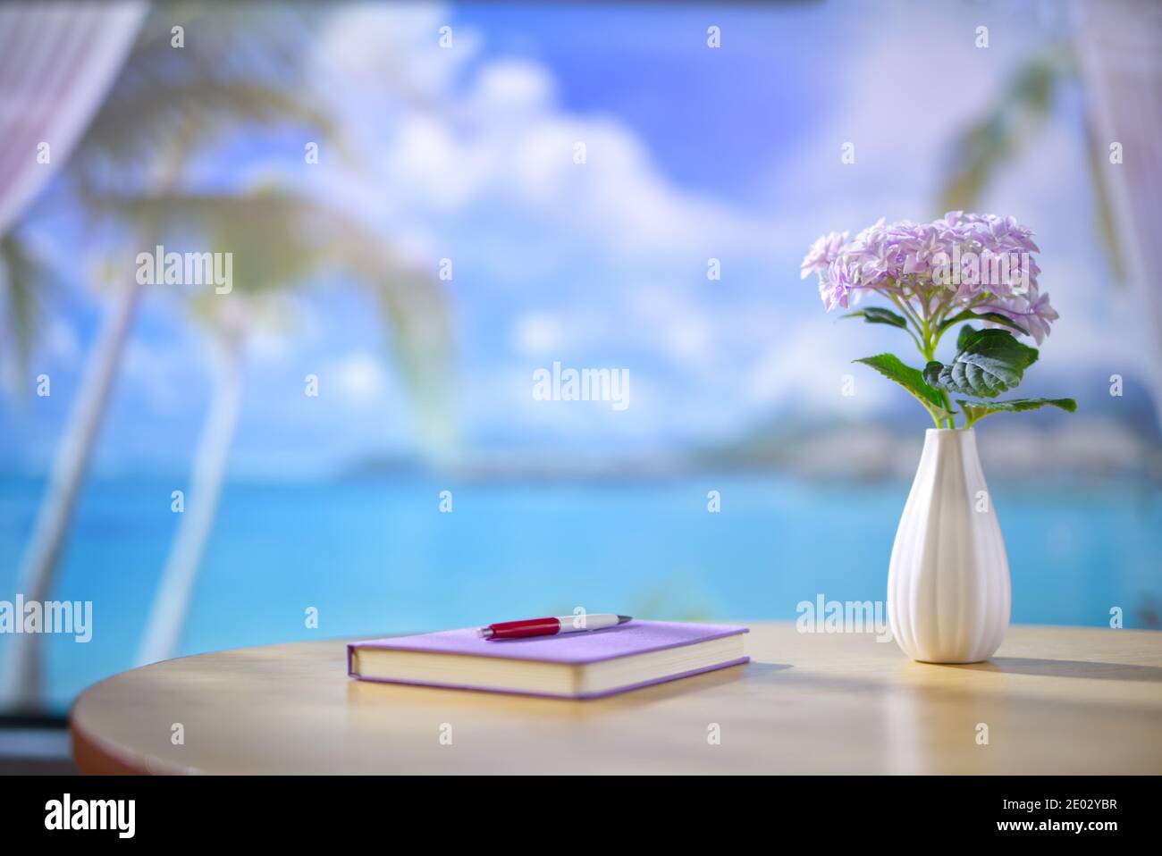 Maison en bord de mer, bureau en bois avec carnet, stylo et vase de fleurs violettes. Belle vue floue du paysage de plage avec ciel bleu et blanc Banque D'Images