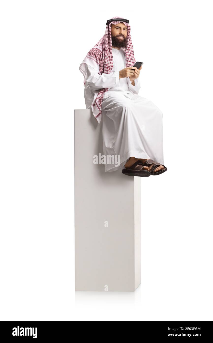 Homme arabe saoudien tenant un smartphone et assis sur un colonne blanche haute isolée sur fond blanc Banque D'Images