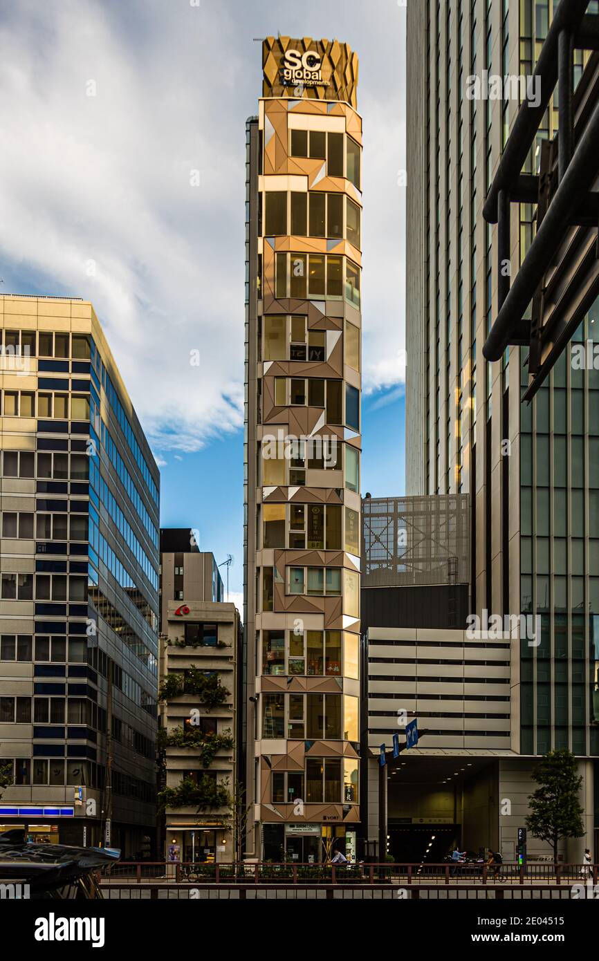Le gratte-ciel étroit « SC Global », sa façade triangulaire dorée captant une lumière chaude, se dresse parmi les tours de bureaux sur Harumi-dori dans le quartier de Chuo, Tokyo, Japon. Banque D'Images