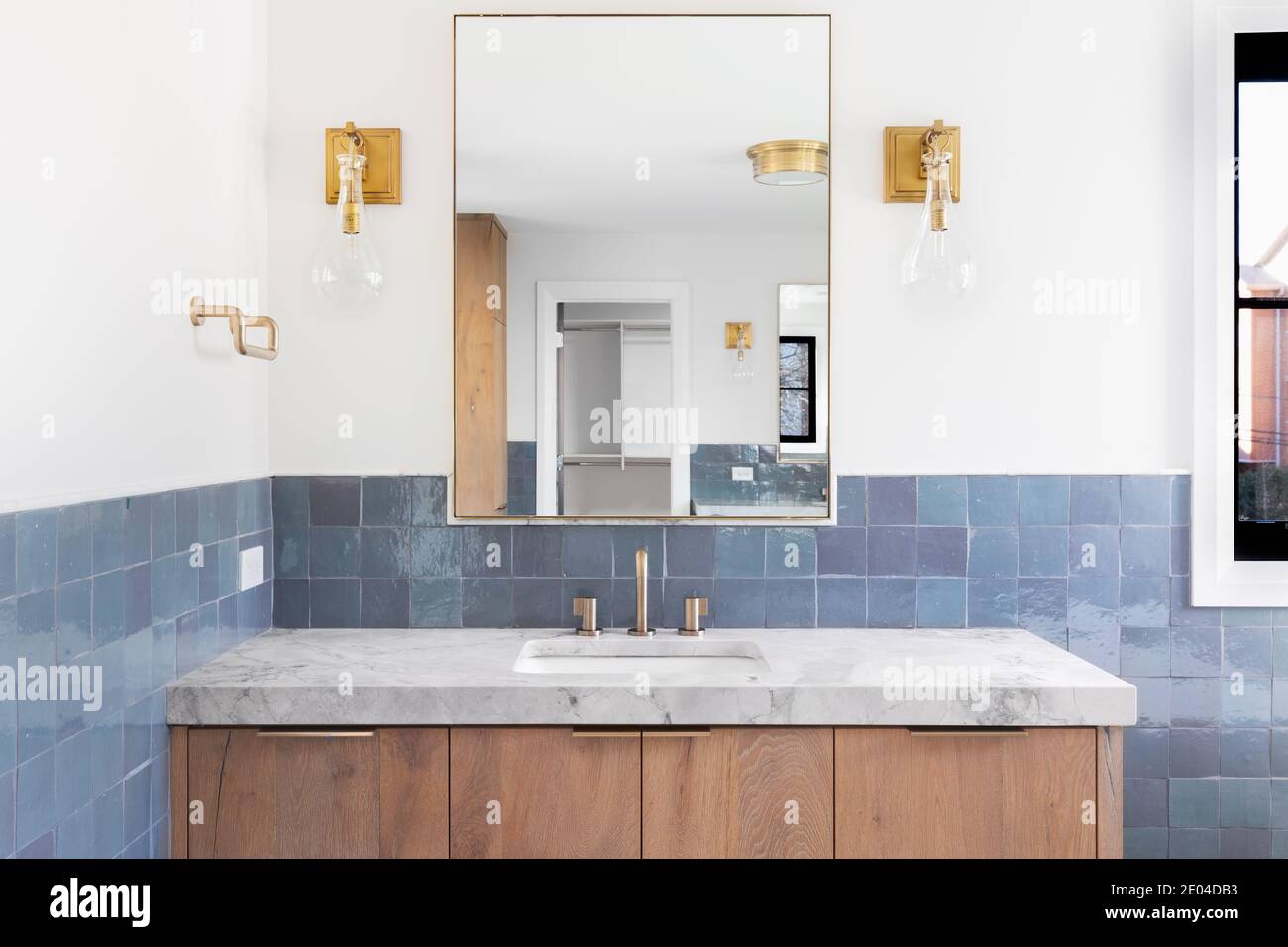 La salle de bains rénovée d'une maison de luxe est dotée de carrelage bleu, d'un meuble-lavabo en bois clair, de lampes dorées montées sur le mur et d'un comptoir en pierre grise. Banque D'Images