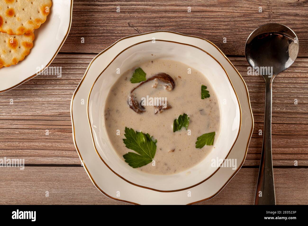 Représentation plate d'une soupe crémeuse aux champignons décorée de persil feuilles dans une tasse en porcelaine artisanale sur une assiette plate sur une table en bois, servi avec un cracker Banque D'Images