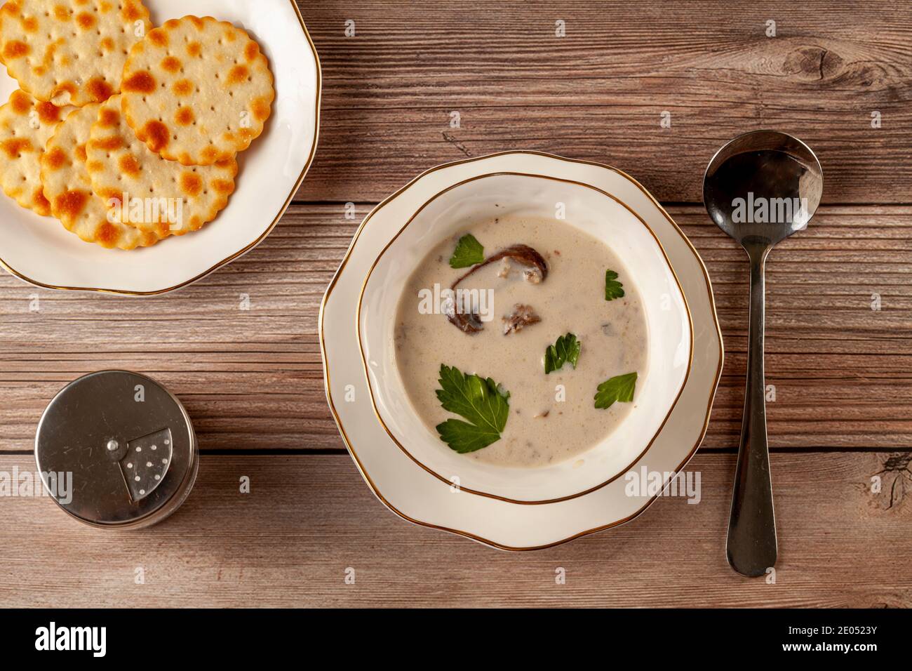 Représentation plate d'une soupe crémeuse aux champignons décorée de persil feuilles dans une tasse en porcelaine artisanale sur une assiette plate sur une table en bois, servi avec un cracker Banque D'Images