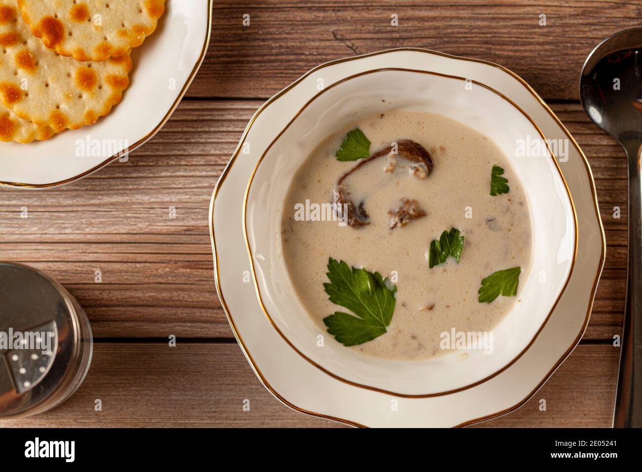 Représentation plate d'une soupe crémeuse aux champignons décorée de persil feuilles dans une tasse en porcelaine artisanale sur une assiette plate sur une table en bois, servi avec un cracker Banque D'Images