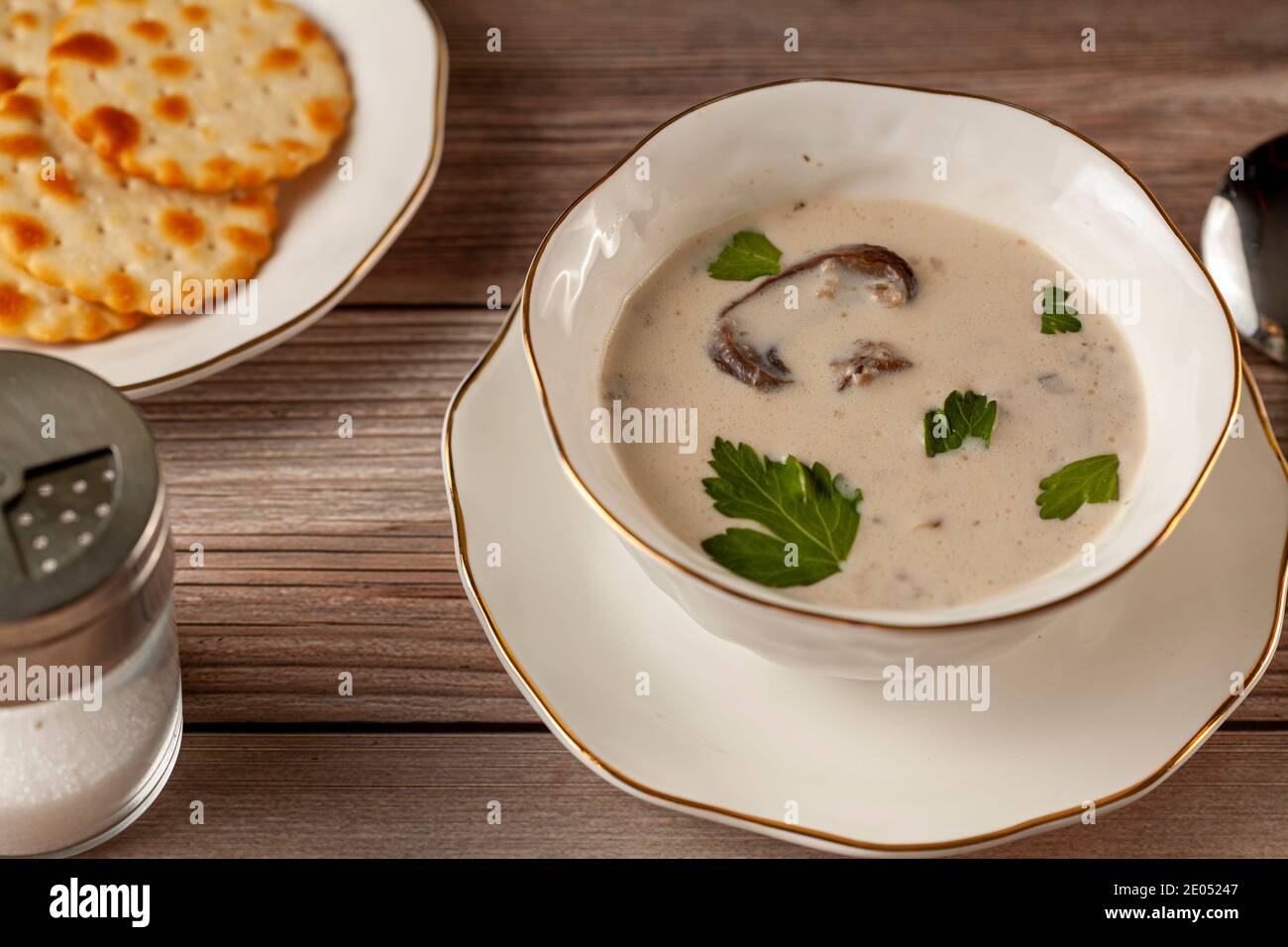 Vue latérale d'une soupe crémeuse aux champignons décorée de feuilles de persil dans une tasse en porcelaine artisanale sur une assiette plate sur une table en bois servie avec des craquelins, Banque D'Images