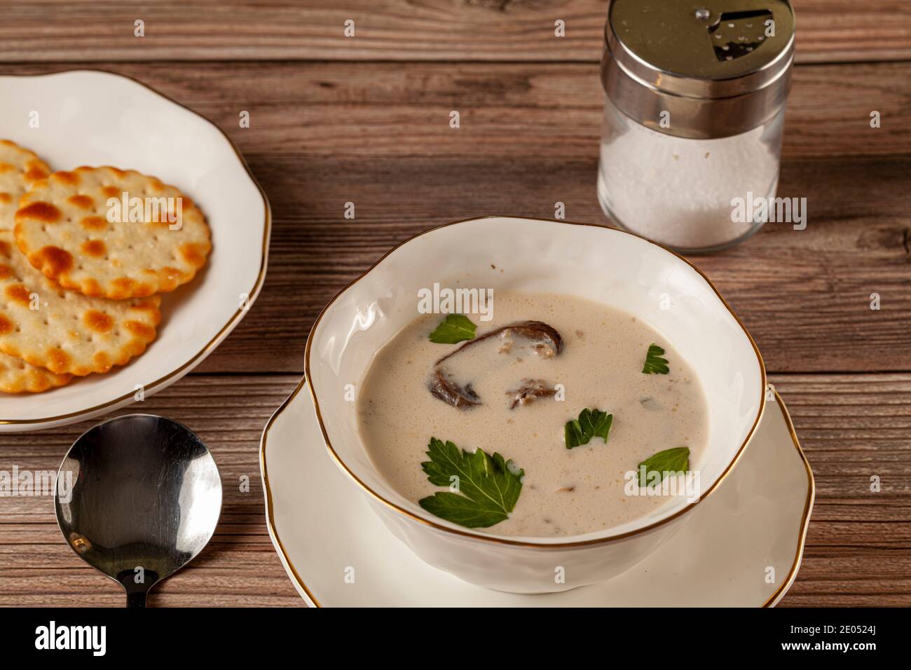 Vue latérale d'une soupe crémeuse aux champignons décorée de feuilles de persil dans une tasse en porcelaine artisanale sur une assiette plate sur une table en bois servie avec des craquelins, Banque D'Images